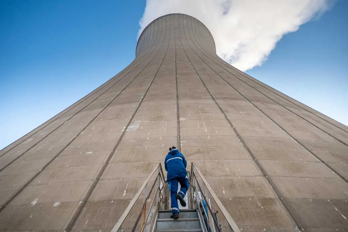 Technicien en combinaison bleue gravissant l'escalier d'une tour de refroidissement nucléaire, contre-plongée