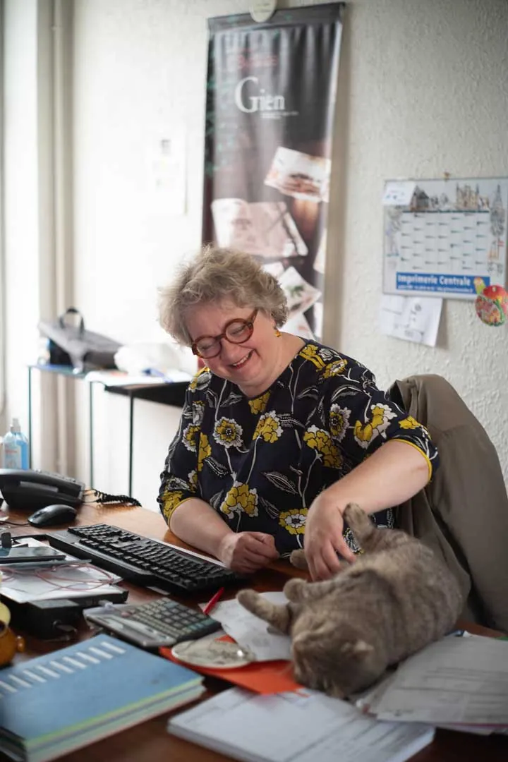 Employée souriante caressant un chat gris sur son bureau, affiche Faïencerie de Gien en arrière-plan