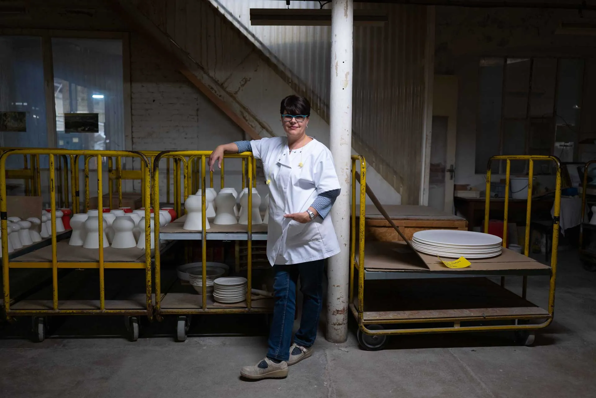 Ouvrière en blouse blanche et lunettes posant accoudée à un chariot de pièces en faïence, Faïencerie de Gien