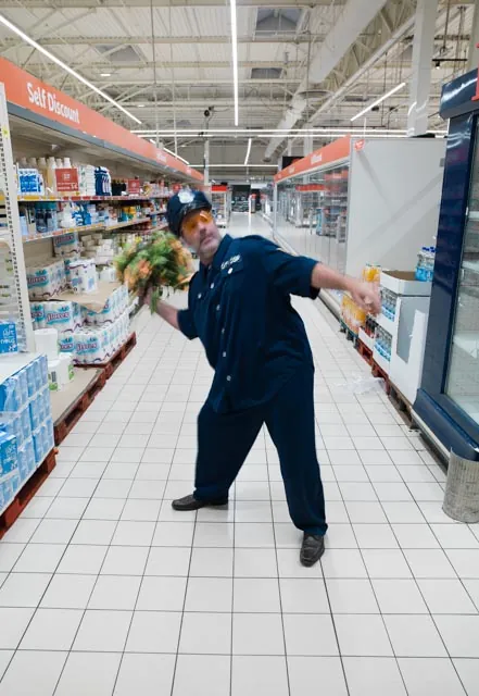Homme en combinaison bleue brandissant un bouquet de fleurs dans une all&eacute;e de supermarch&eacute; d&eacute;sert