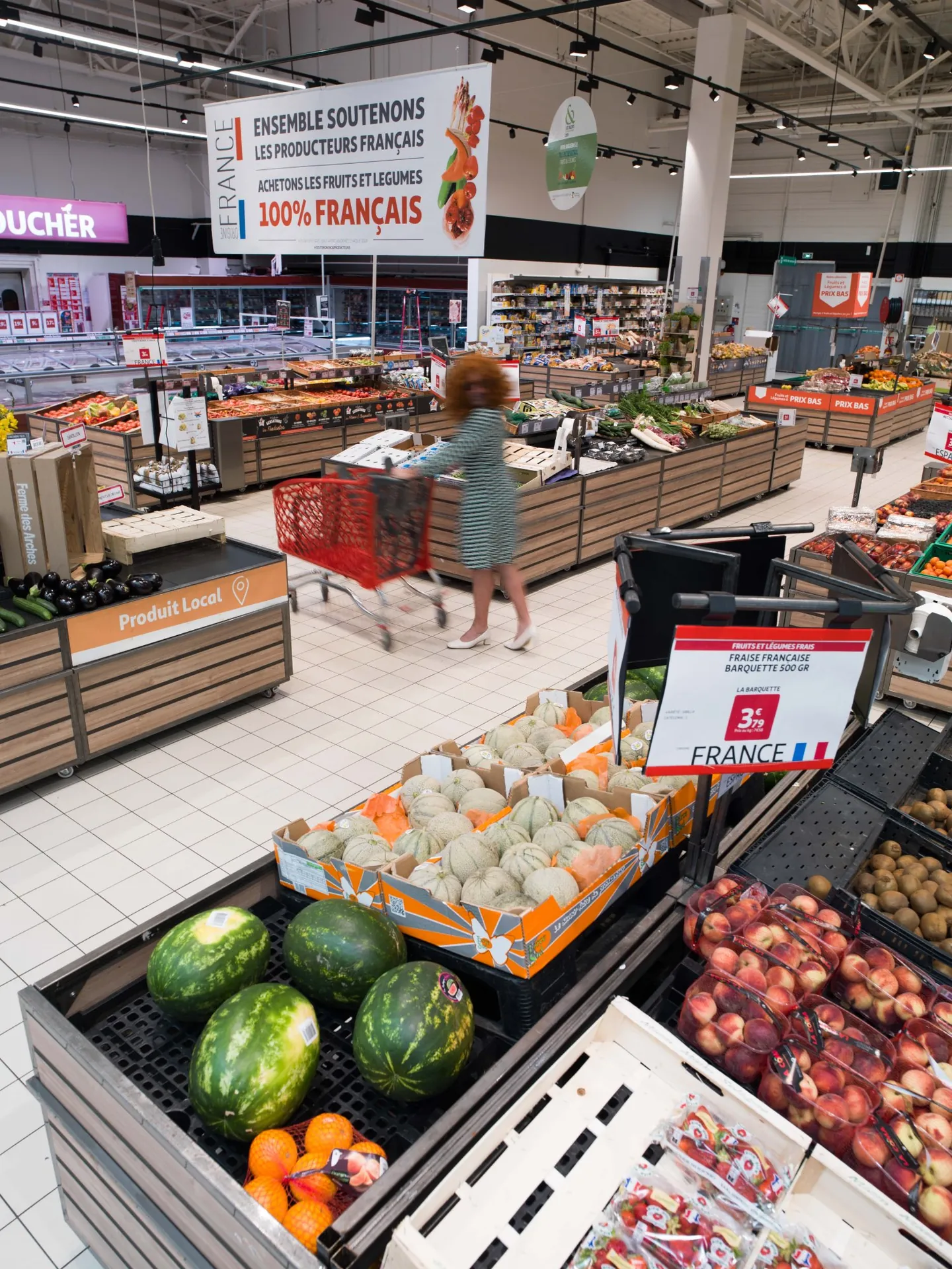 Rayon fruits et l&eacute;gumes, cliente floue avec caddie rouge parmi past&egrave;ques et melons