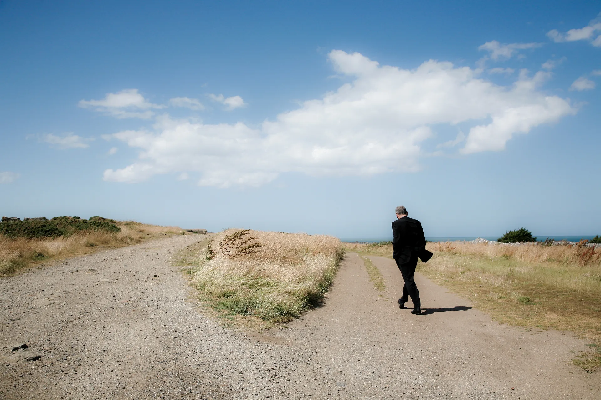 Homme en costume noir marchant de dos sur un chemin côtier vers la mer