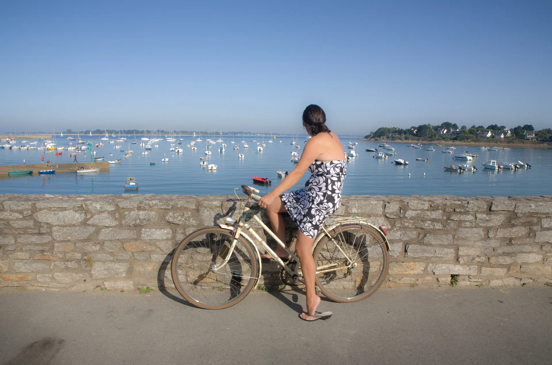Femme à vélo regardant un port avec bateaux, muret en pierre bretonne